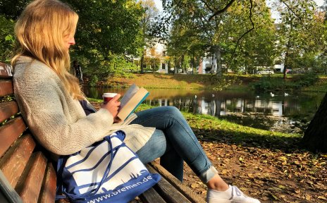 Eine junge Frau sitzt in den herbstlichen Wall-Anlagen auf einer Parkbank, liest ein Buch und trinkt einen Kaffee