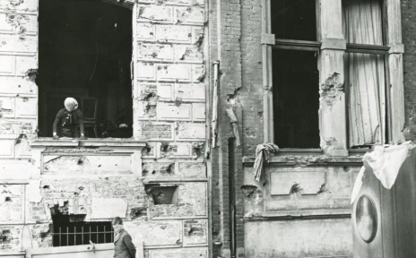 Schwarzweißfoto eines kriegsbeschädigten Hauses mit zerstörter Fassade; ein Kind steht unten vor dem Gebäude, eine Person blickt aus einem Fenster, Trümmer liegen im Vordergrund.