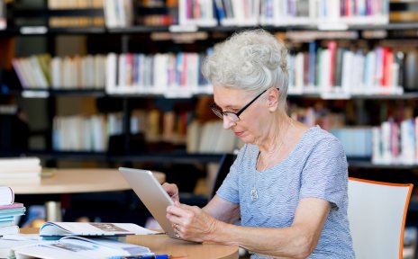 Eine ältere Dame sitzt mit ihrem iPad in der Bibliothek und studiert