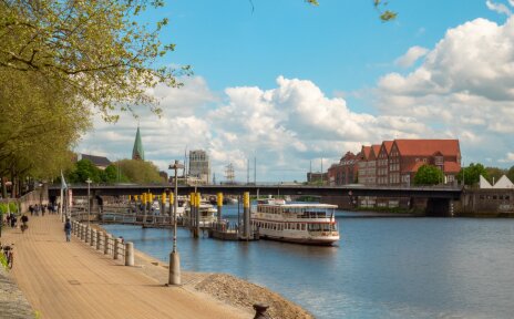Blick die Weserpromenade entlang, am rechten Ufer ist die Weserburg zu sehen, auf dem Wasser einige Schiffe, am linken Ufer Passantinnen und Passanten sowie grün bewachsene Bäume.
