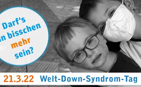 Zwei Jungen umarmen einander, sie schauen glücklich in die Kamera. Auf dem Bild ist der Schriftzug: "Darf's ein bisschen mehr sein? 21.3.22 Welt-Down-Syndrom-Tag".