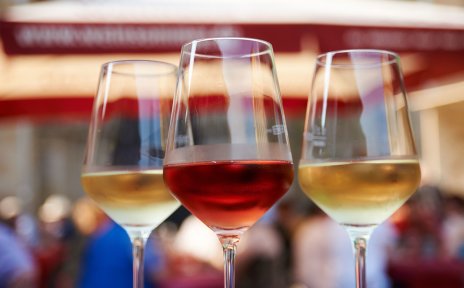 Zwei Weingläser mit einem roten und einem weißen Wein gefüllt stehen in einer Nahaufnahme vor dem Festgetümmel im Hintergrund auf einem Tisch.