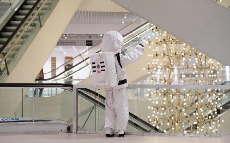 Ein Mensch in Astronautenkleidung steht winkend an einem Geländer. Im Hintergrund sind die Rolltreppen des ehemaligen Kaufhauses zu sehen. 
