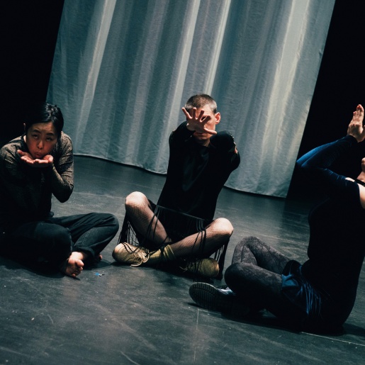 Drei Personen sitzen auf einem schwarzen Boden und machen unterschiedliche Gesten.
