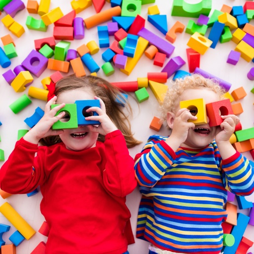 Zwei Kinder halten sich Spielholzklötze vor die Augen und gucken in die Kamera. Um sie herum liegen viele bunte Bauklötze