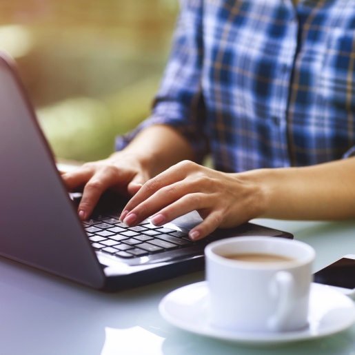Eine Frau tippt auf einer Laptoptastatur, daneben Kaffeetasse und Smartphone.