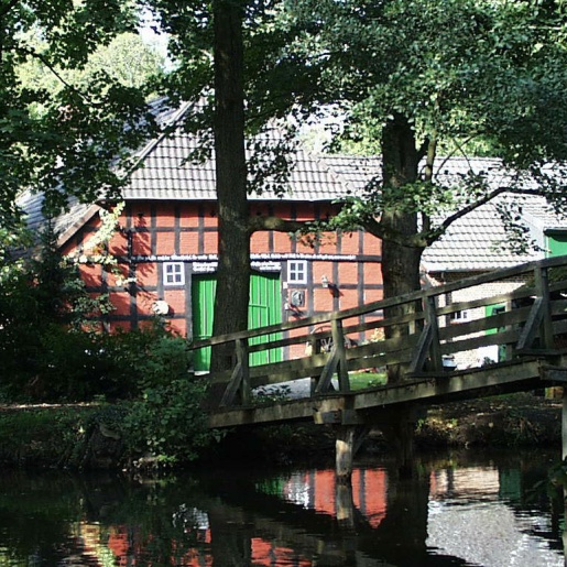 Eine hölzerne Brücke führt über einen Fluss zu einem Fachwerkhaus.