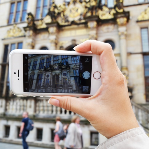 Zu sehen ist eine Hand, die ein Handy hält. Auf dem Handybildschirm ist der Ausschnitt eines Gebäudes zu erkennen. Dieses Gebäude sieht man auch im Hintergrund des gesamten Bildes. Es hat eine goldene Fassade und Treppen.