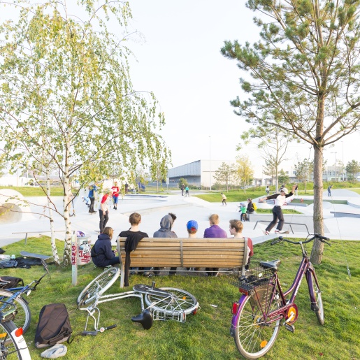 Eine Holzbank von hinten, auf der Personen sitzen mit Blick auf die Rampen eines Skateparks. Drumherum liegen zahlreiche Fahrräder im Gras.