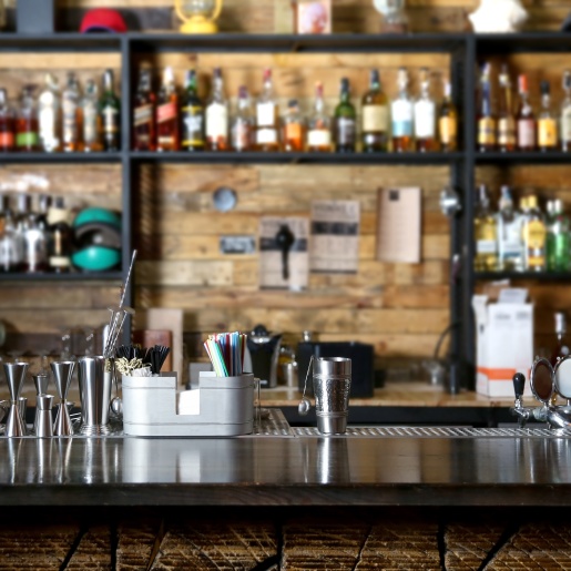 Eine Theke ist im Zentrum des Bildes. Am Rand steht ein Barkeeper. Im Regal sind verschiedene Flaschen und Utensilien zum Cocktails mixen. 