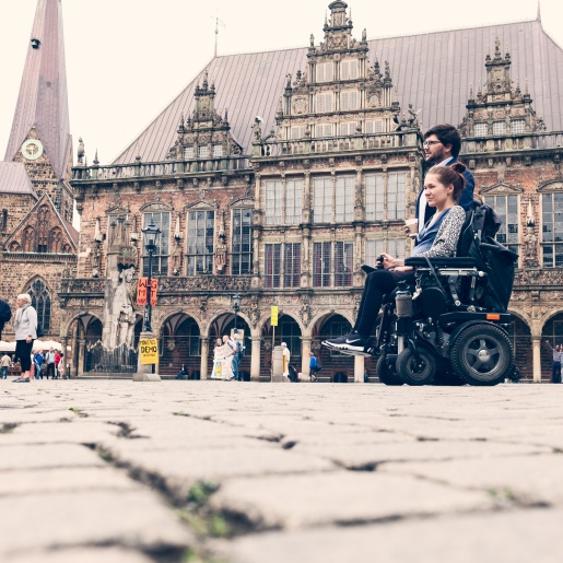 Ein Mann und eine Frau auf dem Marktplatz vor dem Bremer Rathaus. Die Frau sitzt im Rollstuhl.