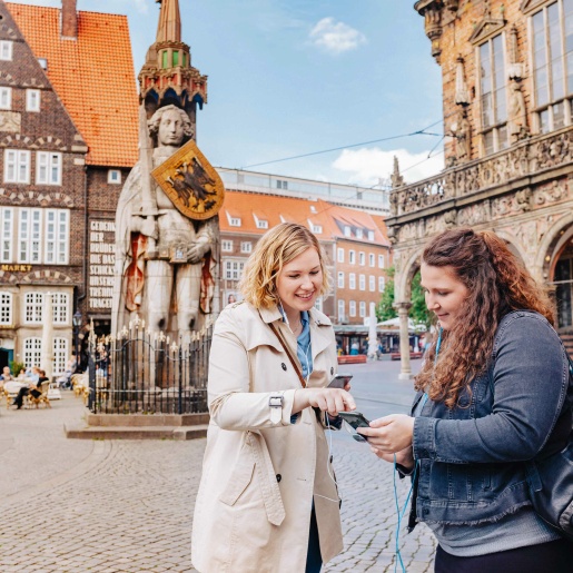Zwei Frauen gucken sich auf dem Bremer Marktplatz den Audioguide an und haben Kopfhörer im Ohr.