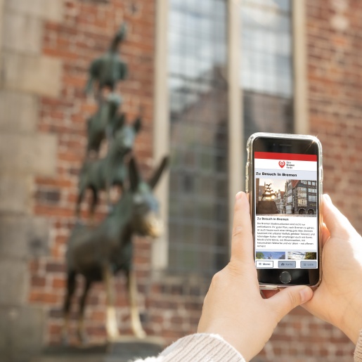 Jemand hält ein Smartphone vor den Stadtmusikanten. Auf dem Bildschirm sieht man die Startseite von "Dein Bremen Guide".