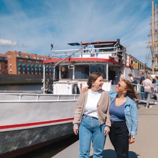 Zwei Frauen lachen sich an. Im Hintergrund gehen Leute an Bord eines Schiffes.