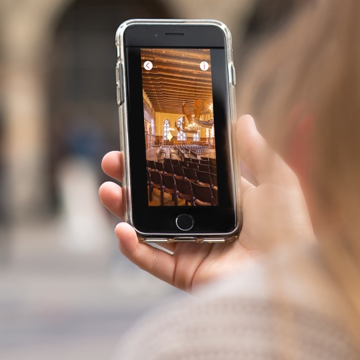 Eine Frau hält ein Smartphone in der Hand. Darauf sieht sie ein Bild vom Inneren des Bremer Rathauses. 