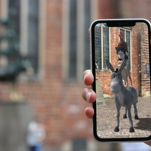 Im Vordergrund sind die Bremer Stadtmusikanten in 3D durch ein Smartphone zu sehen. Im Hintergrund ist die Statue der Stadtmusikanten zu sehen. 
