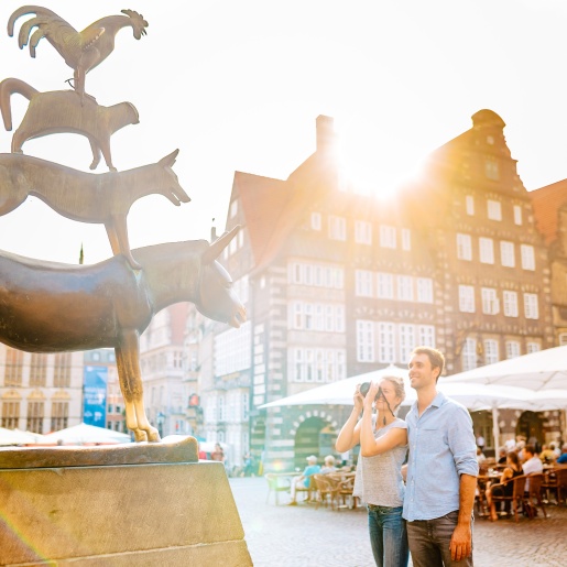 A woman and a man stand in front of the Bremen Town Musicians. The woman takes a picture of the bronze statue.