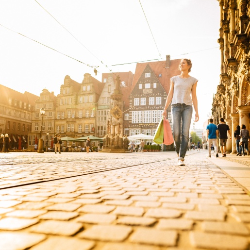 A woman walks across a square with shopping bags. Historical buildings can be seen in the background. The sun is shining.