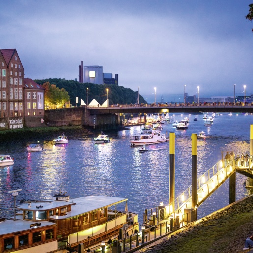 Auf dem Fluss "Weser" sind mehrere beleuchtete Schiffe zu sehen. Einige Menschen stehen auf einer Brücke und schauen den Schiffen zu. Es ist Abenddämmerung.