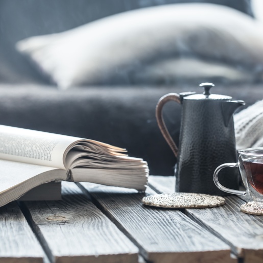 Ein aufgeschlagenes Buch liegt auf einem Holztisch, daneben steht eine Teekanne und ein gefülltes Teeglas. Im Hintergrund ein Sofa mit Kissen.