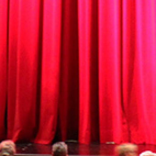 Ein großer roter geschlossener Vorhang, vor dem einige Menschen sitzen.