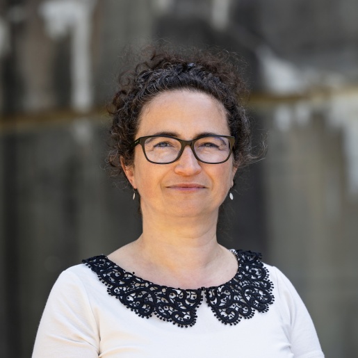 Eine Frau in einem weißen Tshirt, schwarzer Brille und hochgebundenen schwarzen Haaren lacht. 