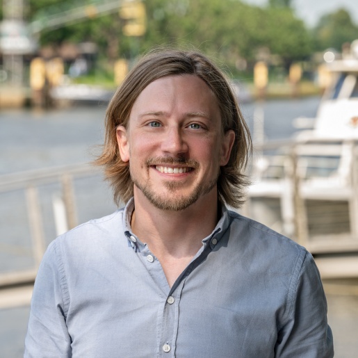 Ein Mann lächelt freundlich in die Kamera. Hinter ihm fließt ein Fluss.