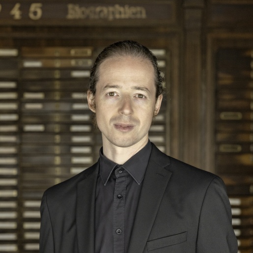 Portrait von einem schlanken Mann mit zurück gebundenen Haaren. Hinter ihm ist eine Wand aus Holz zu erkennen