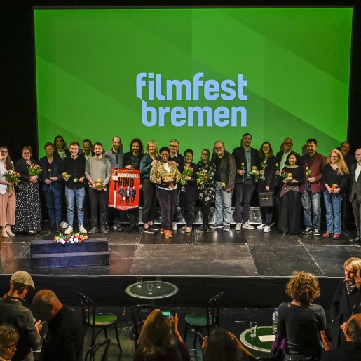 Eine Gruppe von Menschen steht auf einer Bühne vor einer Leinwand, auf der "Filmfest Bremen" geschrieben steht.