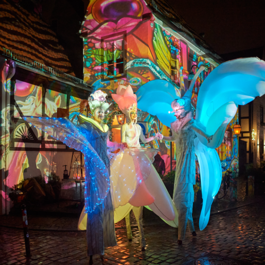 Costumed stilt walkers stand in front of the colourfully illuminated house facades in the Schnoor.