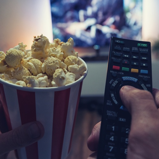 Eine Person sitzt vor einem Fernseher und hält eine Fernbedienung und einen Becher Popcorn in der Hand