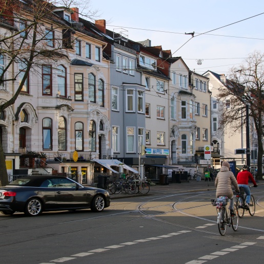 Zwei Fahrradfahrer und ein Auto fahren über eine Kreuzung. Im Hintergrund reihen sich Altbremer Häuser aneinander. 