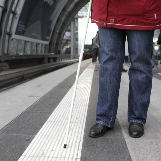 Eine Person steht auf einem Bahnsteig und ertastet mit einem Langstock das Bodenleitsystem. 