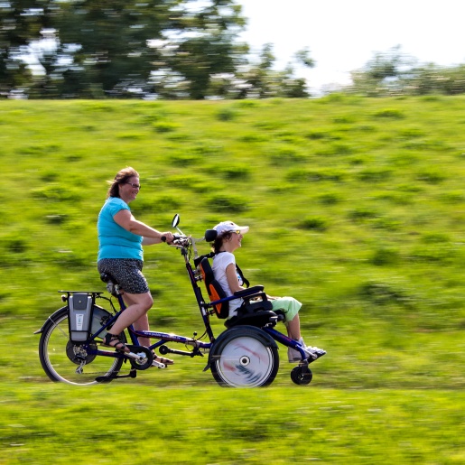 Zwei Menschen rasen mit einem Fahrrad durchs Grüne. Eine Person lenkt, die andere sitzt in einem Sitz vor dem Lenkrad.