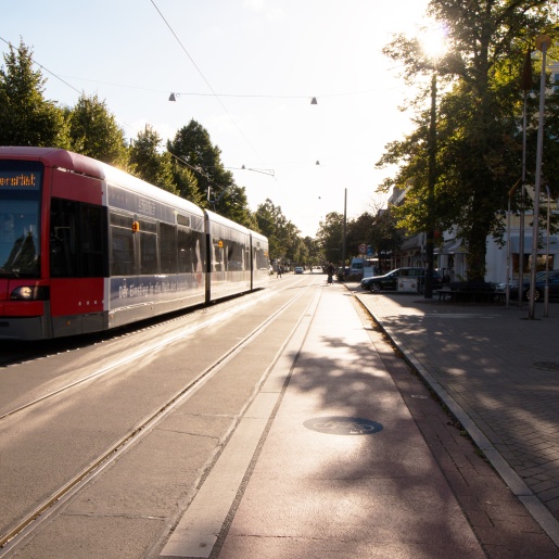 Blick in die Wachmannstraße. Eine Straßenbahn fährt vorbei. Im Hintergrund sind Autos und Personen, die die Fahrbahn überqueren. Außerdem ist eine Person auf einem Dreirad zu sehen.
