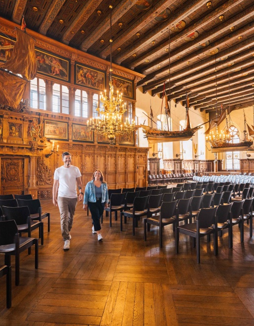 Zwei Personen laufen durch einen historischen Saal mit Stühlen. Schiffe hängen von der Decke.