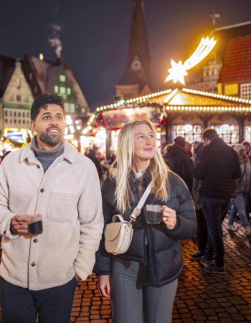 Ein Mann und eine Frau laufen mit Tassen in der Hand über den Bremer Weihnachtsmarkt.