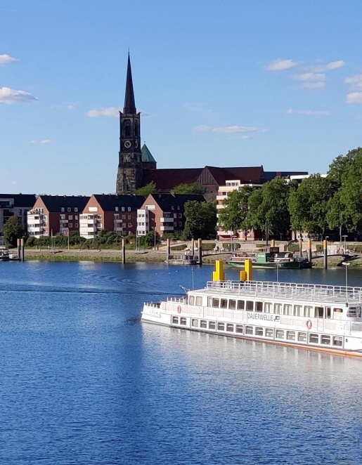 Ein weißes Schiff mit dem Namen "Dauerwelle" liegt an einem Anleger vor dem Teerhof. Im Hintergrund die verlängerte Schlachte-Promenade, saftig belaubte Bäume und strahlend blauer Himmel.