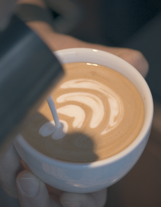 Aus einer Milchkanne wird Milch in eine kleine Tasse mit Kaffee gegossen und ergibt dort ein Muster