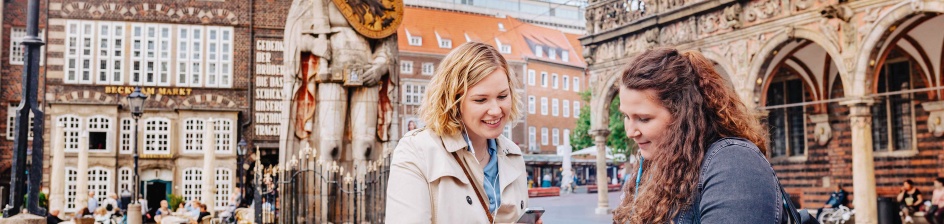 Two women look at the audio guide on Bremen's market square with headphones in their ears.