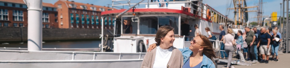 Zwei Frauen lachen sich an. Im Hintergrund gehen Leute an Bord eines Schiffes.