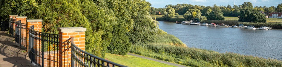Blick über den Knoopspark in Bremen mit geschwungenem Geländer, grünen Uferwiesen und der Lesum im Sonnenschein.