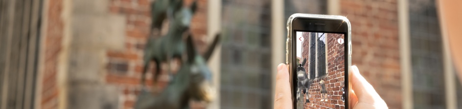 Ein Smartphone mit animierten Stadtmusikanten Figuren wird vor die Statue der Stadtmusikanten gehalten.