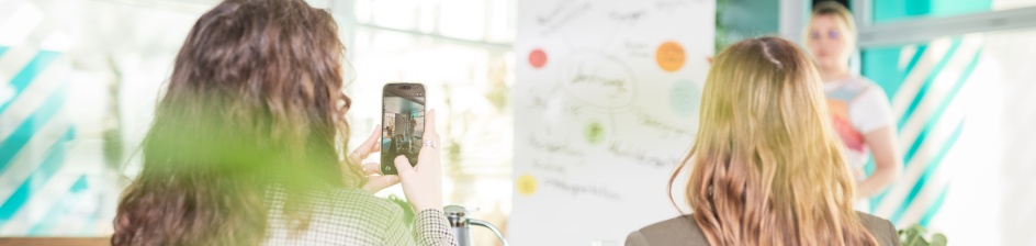 Eine Person trägt etwas an einem Flipchart vor, zwei weitere Personen schauen zu und eine filmt die vortragende Person.