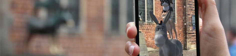 Im Vordergrund sind die Bremer Stadtmusikanten in 3D durch ein Smartphone zu sehen. Im Hintergrund ist die Statue der Stadtmusikanten zu sehen. 