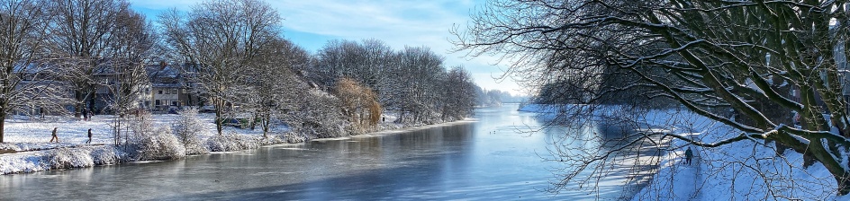 Der Blick geht über ein Gelände auf den Werdersee. Er ist zugefroren. Die Natur am Rand ist mit Schnee bedeckt und man sieht Leute spazieren.