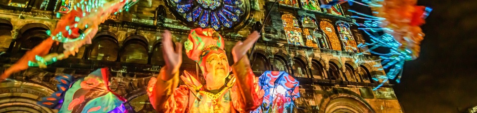 Menschen auf Stelzen und in Verkleidungen stehen vor dem Dom, der im Rahmen vom Lichtertreiben beleuchtet wird.