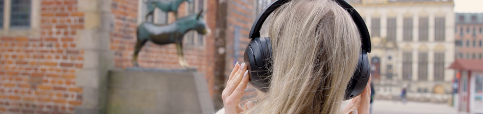Eine Person mit Kopfhörern steht vor den Bremer Stadtmusikanten am Rathaus und hört einen Audiowalk.