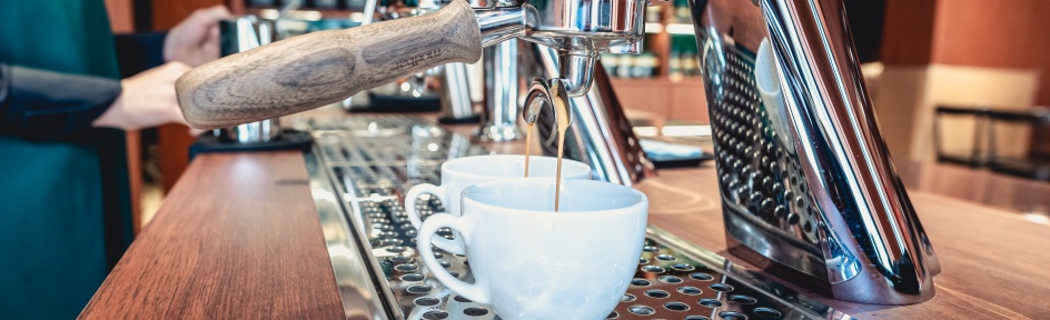 Aus einer silbernen Kaffeemaschine läuft Kaffee in weiße Tassen.