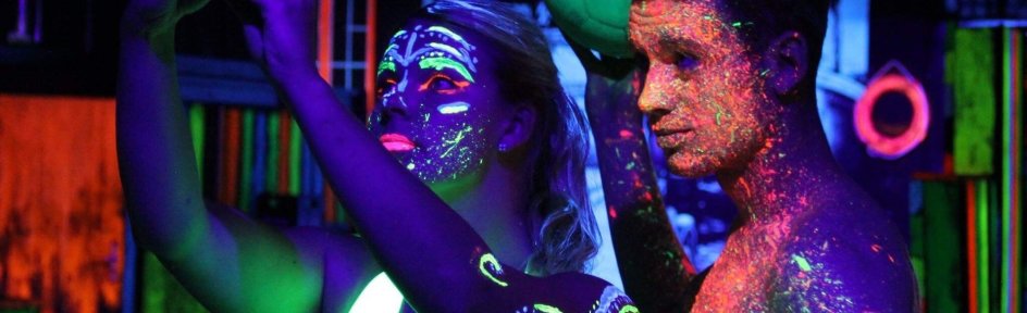 Auf dem Foto sind zwei Jugendliche abgebildet, die im Dunkeln ein Selfie machen. Beide sind mit Neonfarben verschmiert und schauen in die Kamera.
